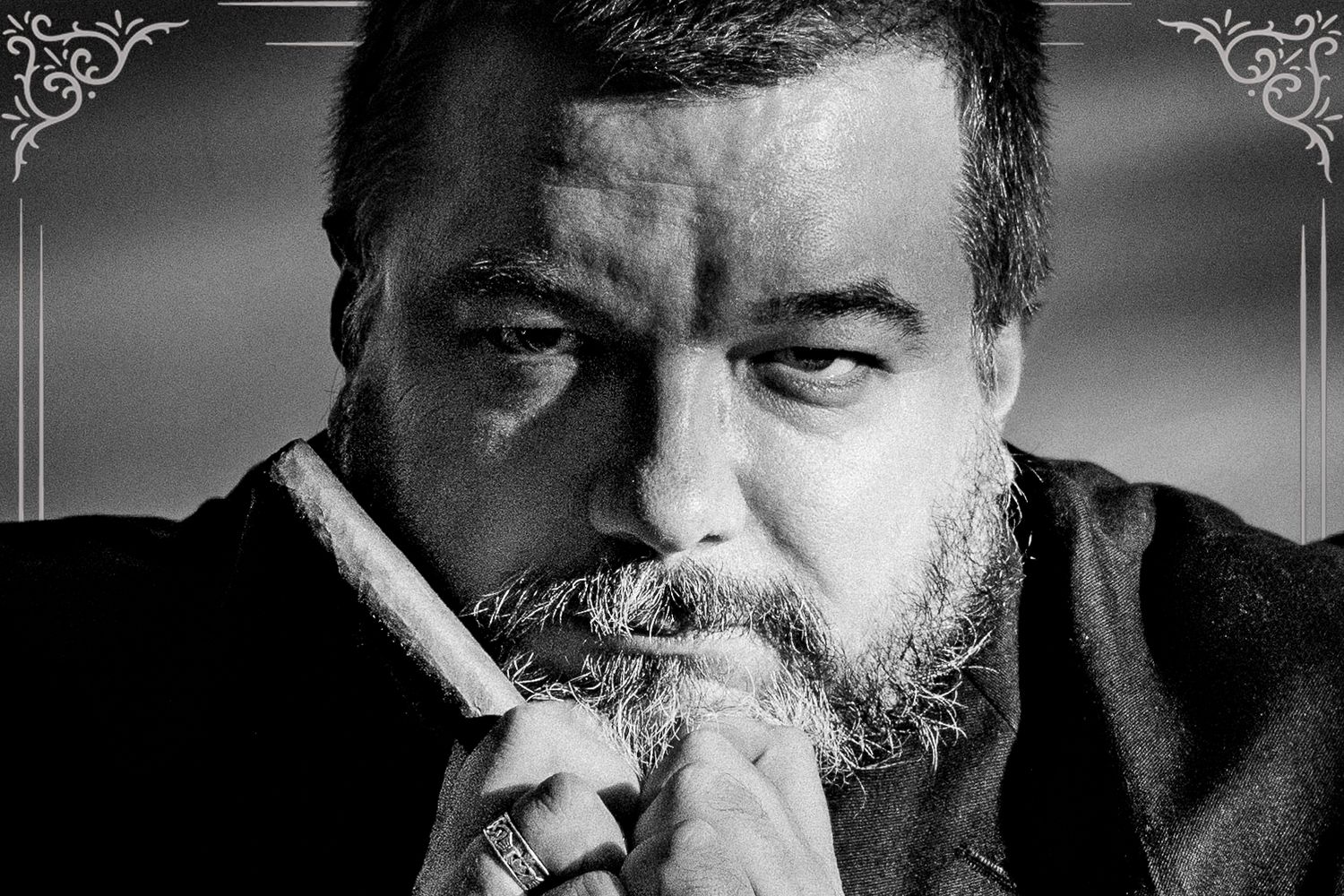 Black and white portrait of a bearded man holding a cigar, looking intensely at the camera.