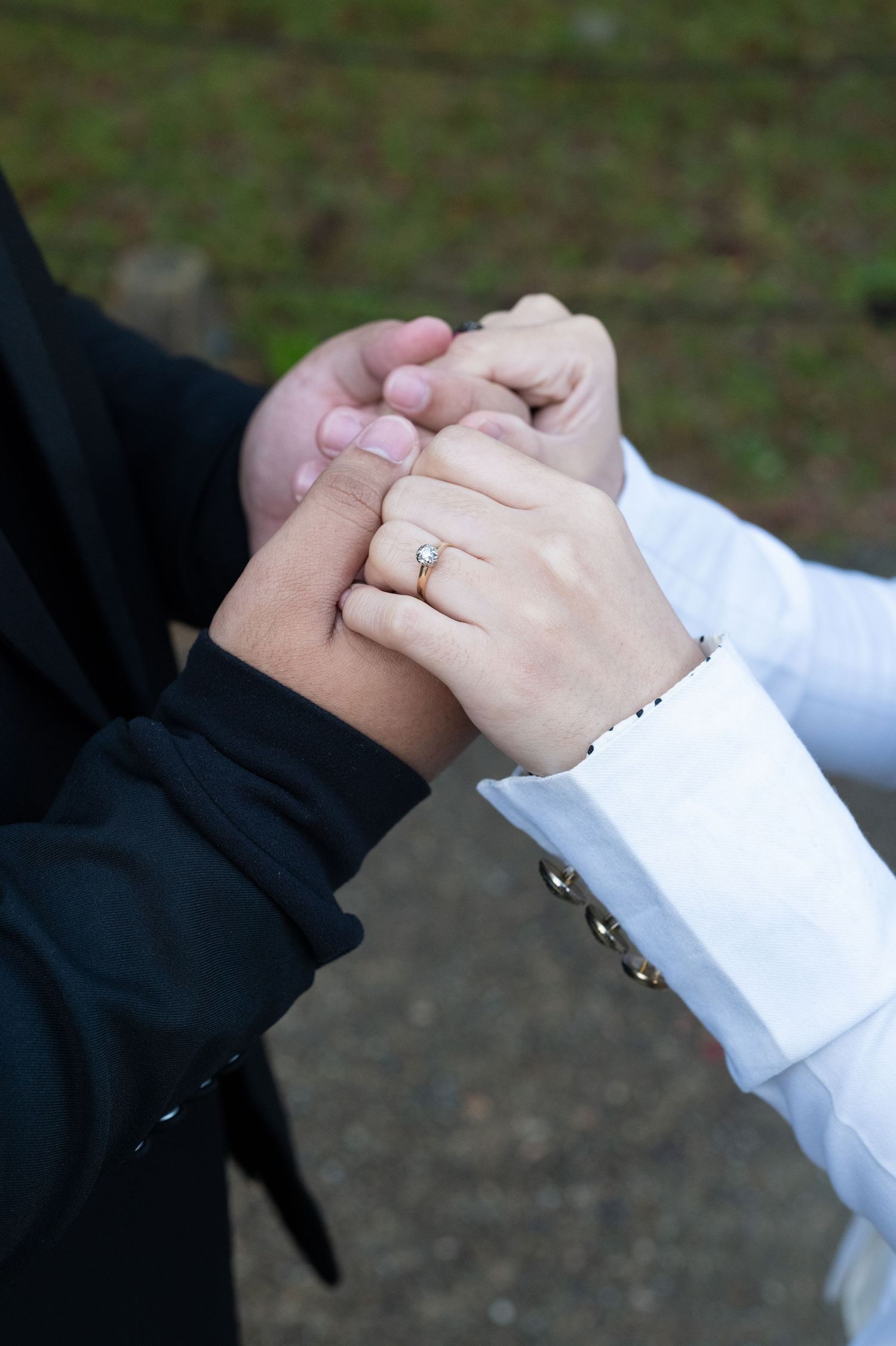Close-up of couple holding hands showing diamond engagement ring in outdoor garden setting.