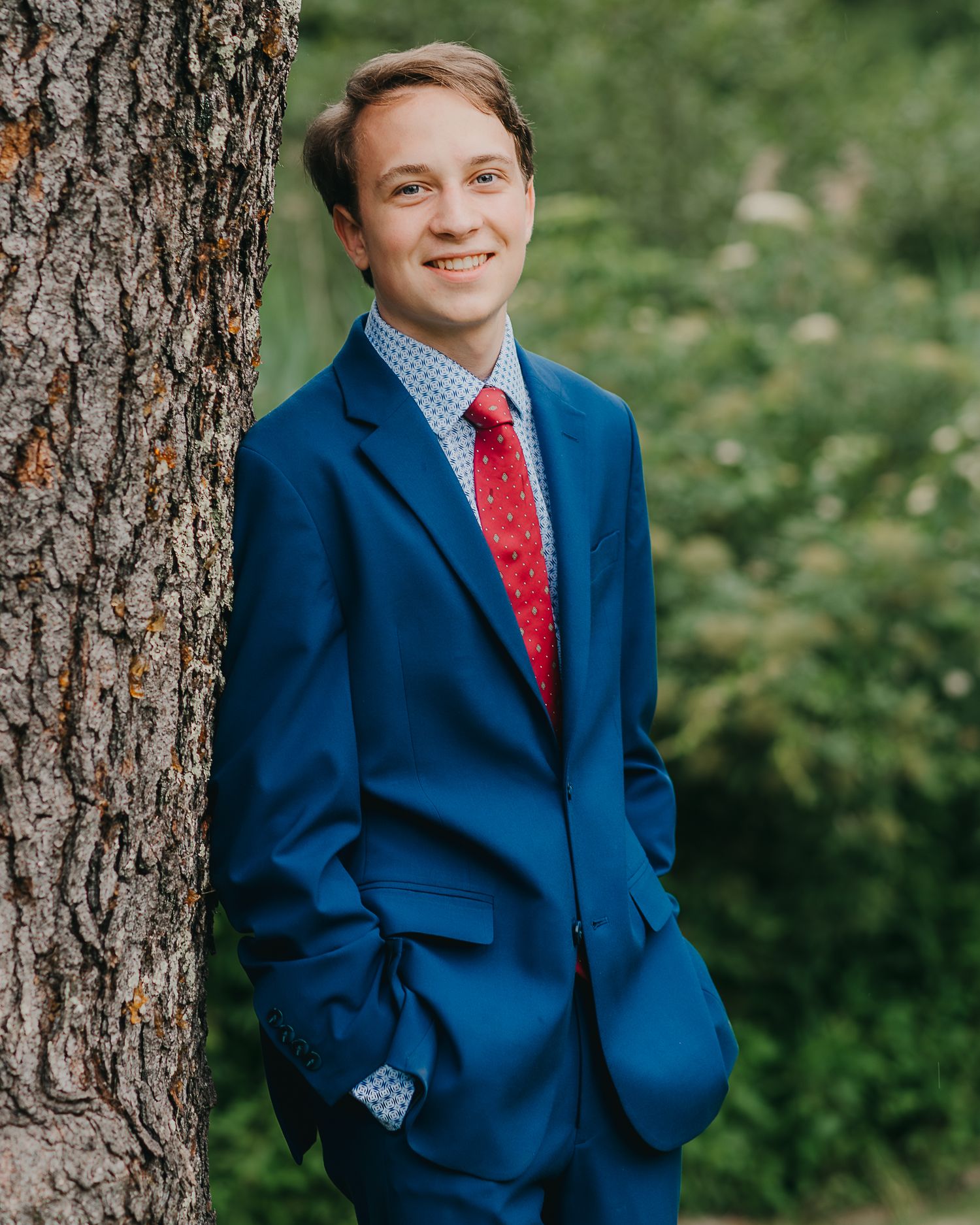 Young man in blue suit and red tie smiling outdoors, leaning against a tree with green foliage background.