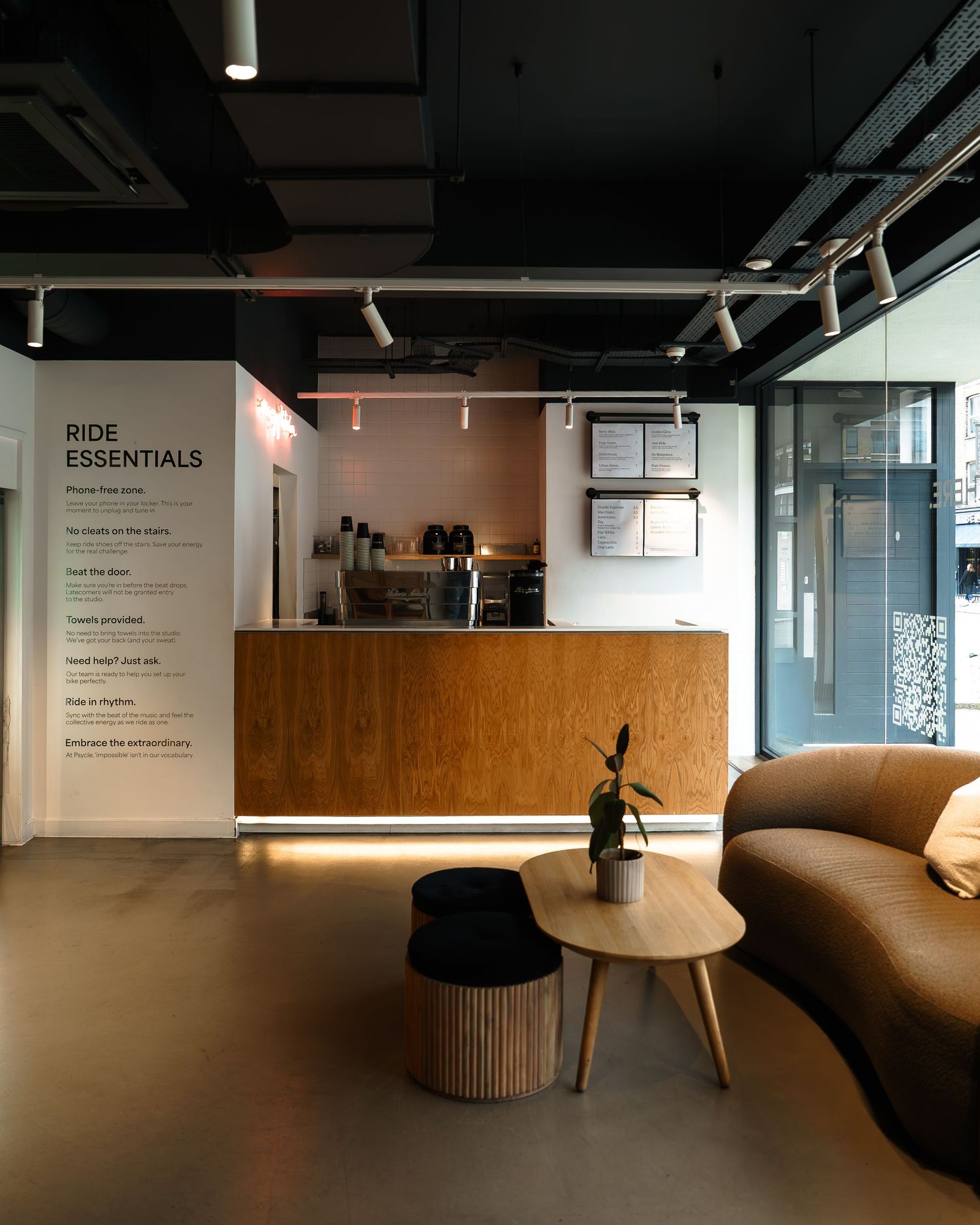 Modern cycling studio lobby with wooden reception desk, brown sofa, round coffee table, and Ride Essentials signage on white wall.