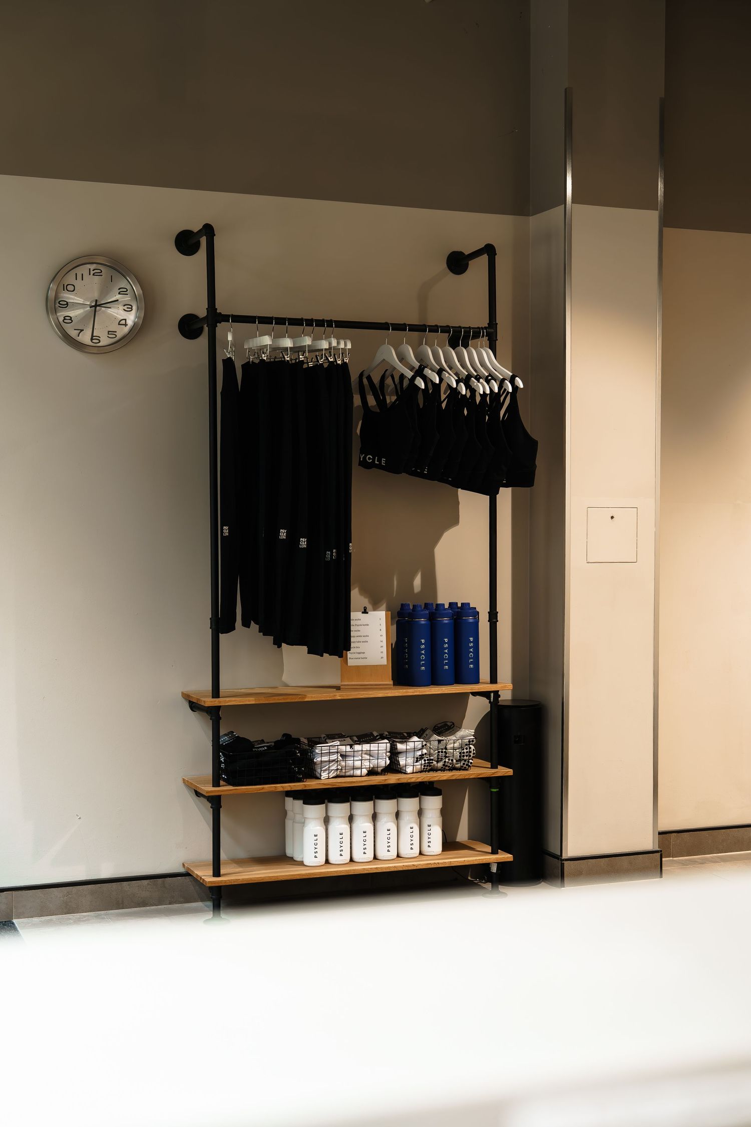 Black clothing rack with wooden shelves displaying folded apparel and blue bottles, beside a wall clock.