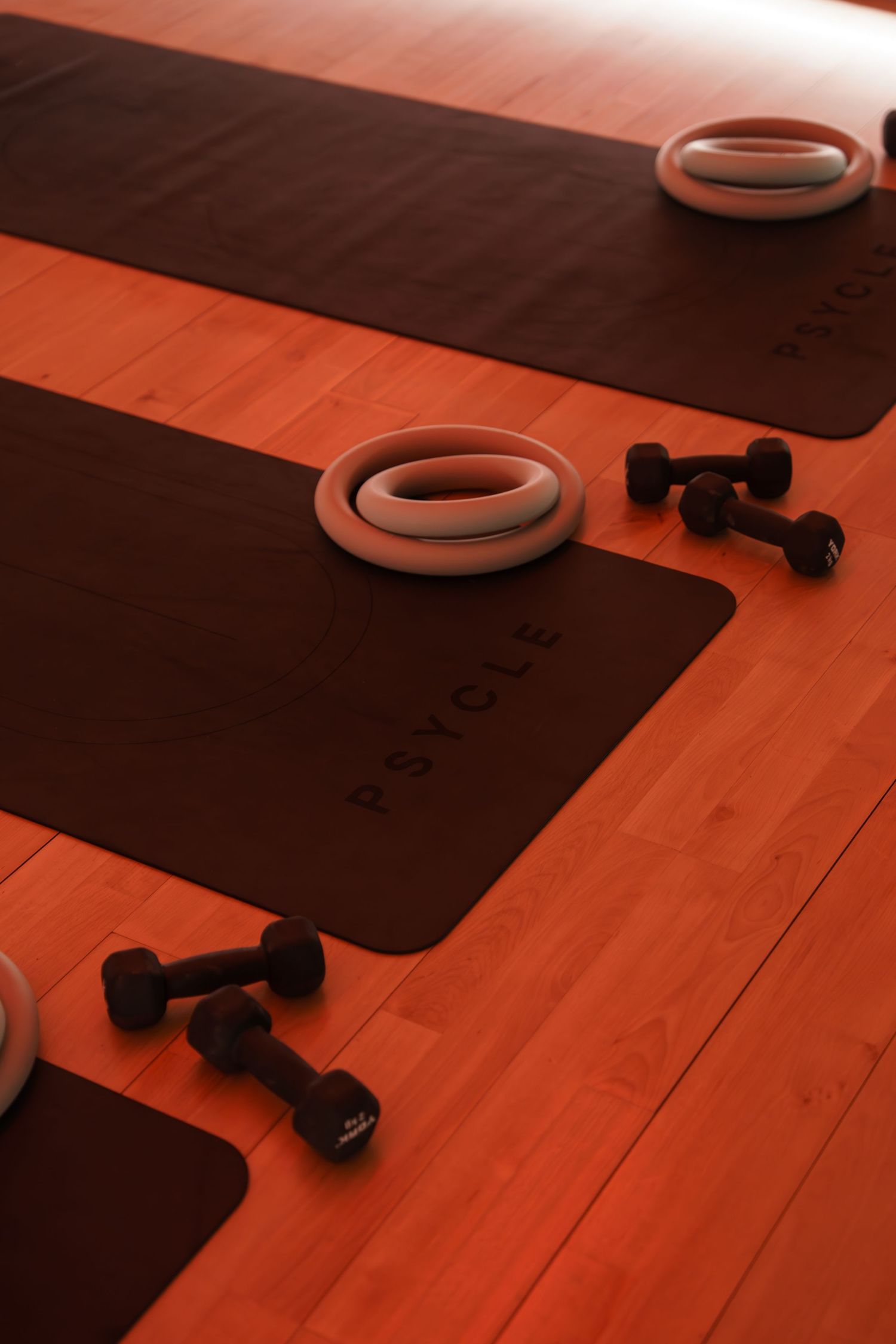 Psycle branded yoga mats with pilates rings and dumbbells on a warm-lit wooden floor in a fitness studio.