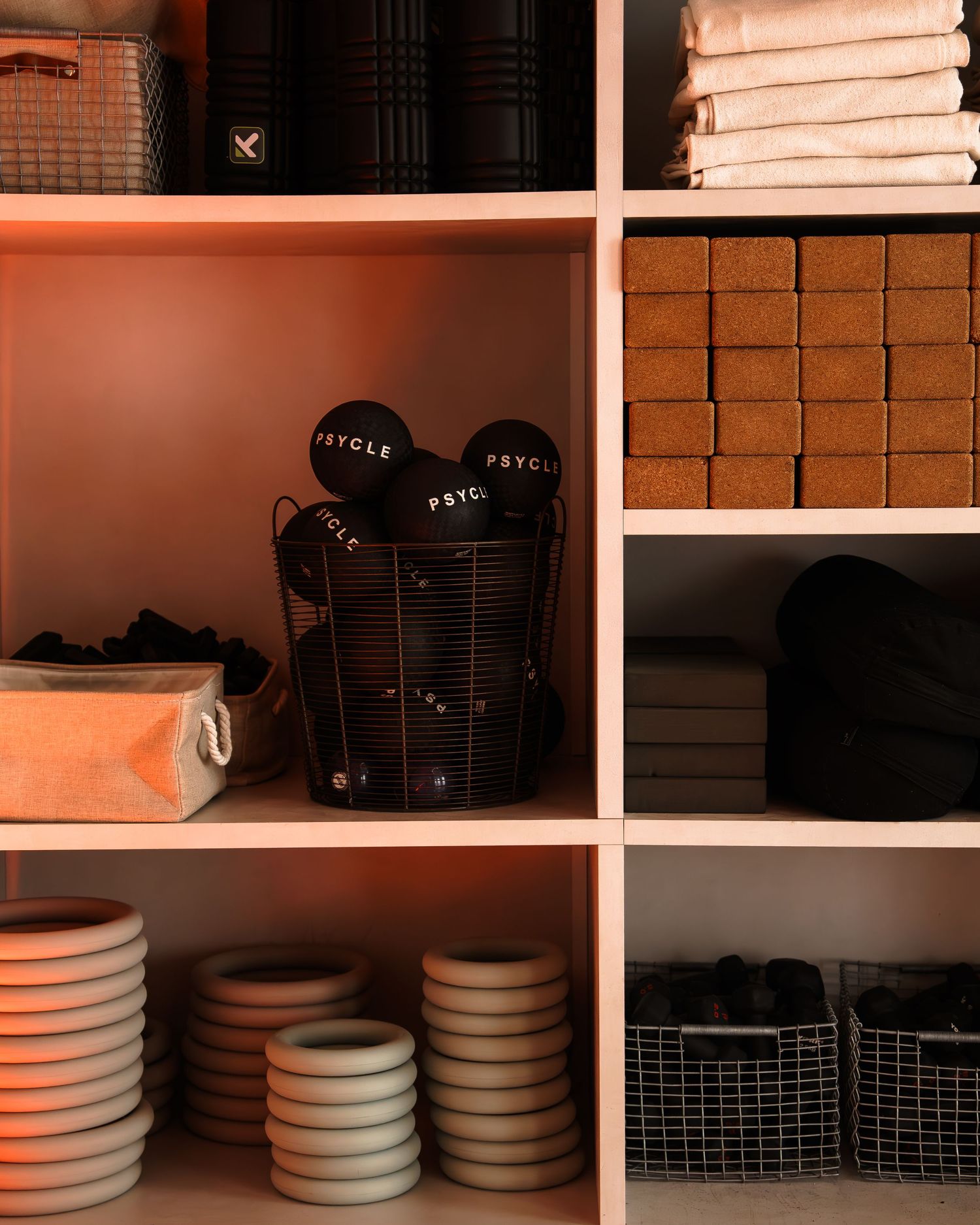 Organized pink shelving unit displaying folded towels, cork blocks, Psycle branded caps in wire basket, and ceramic bowls.