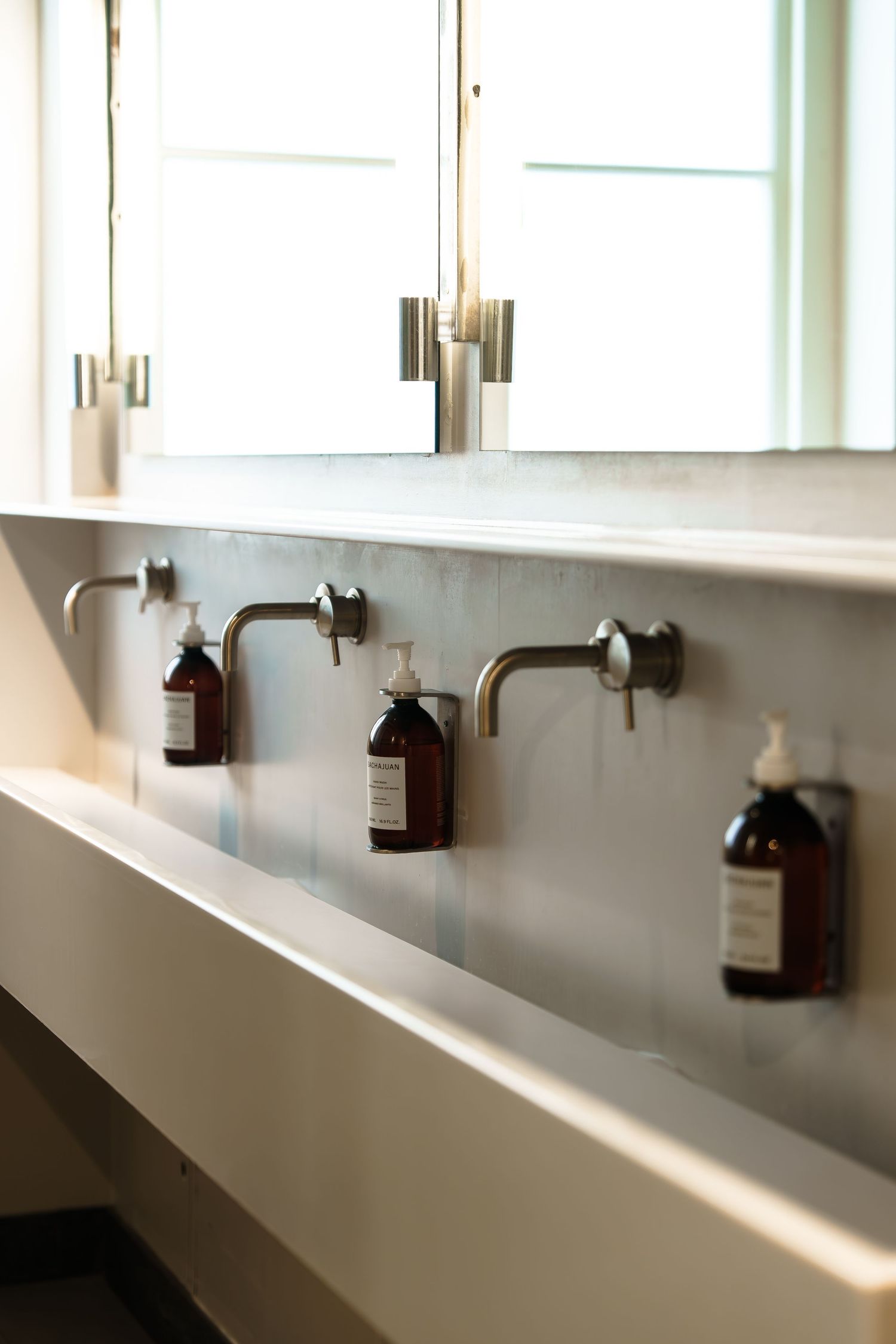 Modern trough sink with wall-mounted faucets and amber soap dispensers beneath a window in a minimalist bathroom.