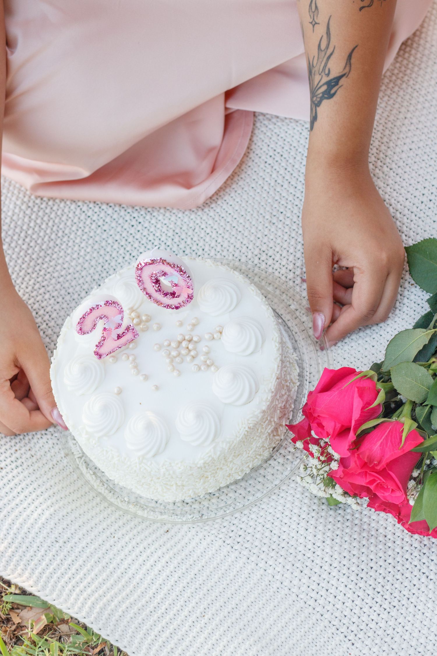 Hands resting beside a white birthday cake topped with number two candles next to pink roses on a blanket.