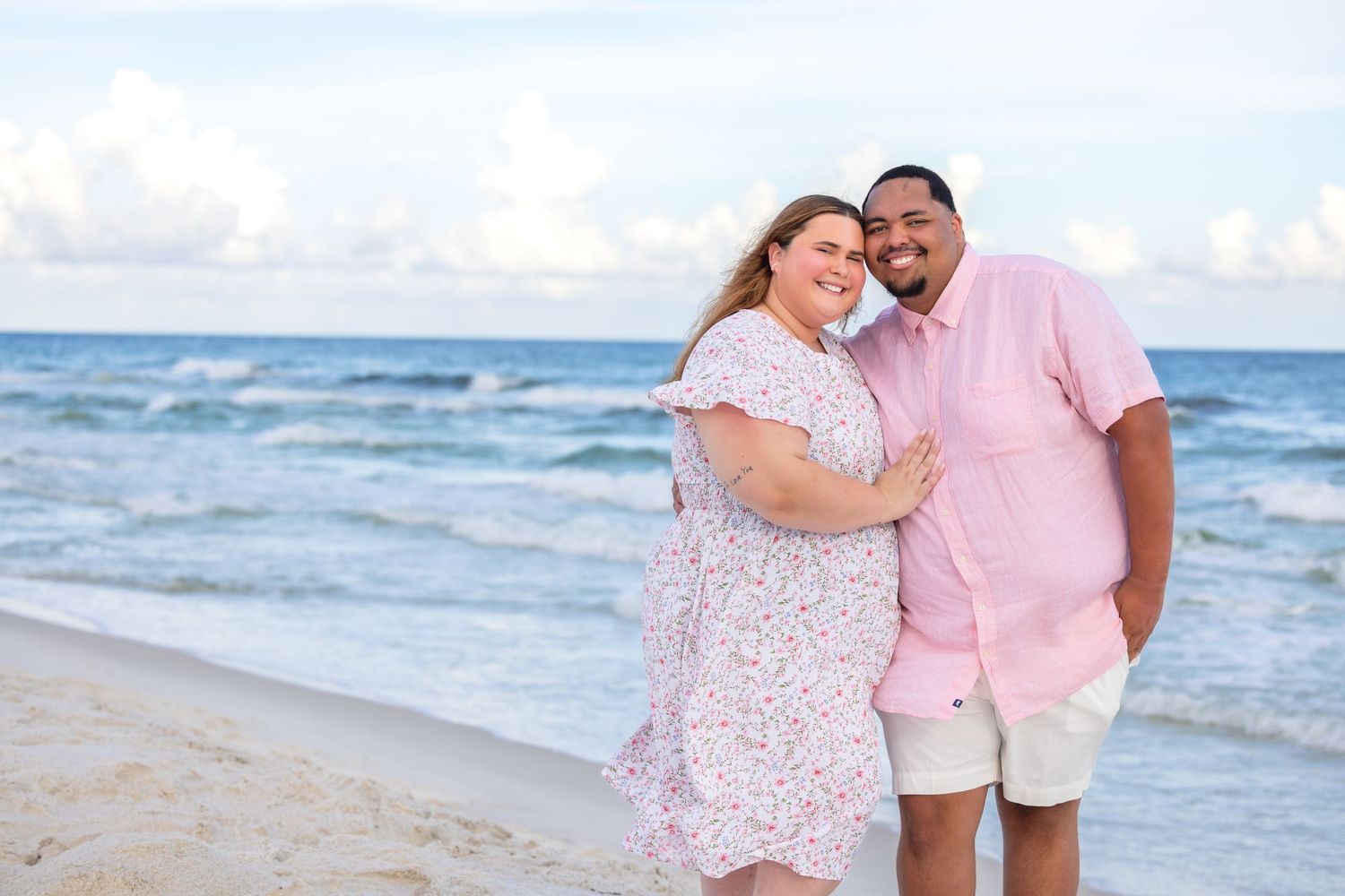 Couple smiling and embracing on a sandy beach, wearing pink outfits with ocean waves in the background.