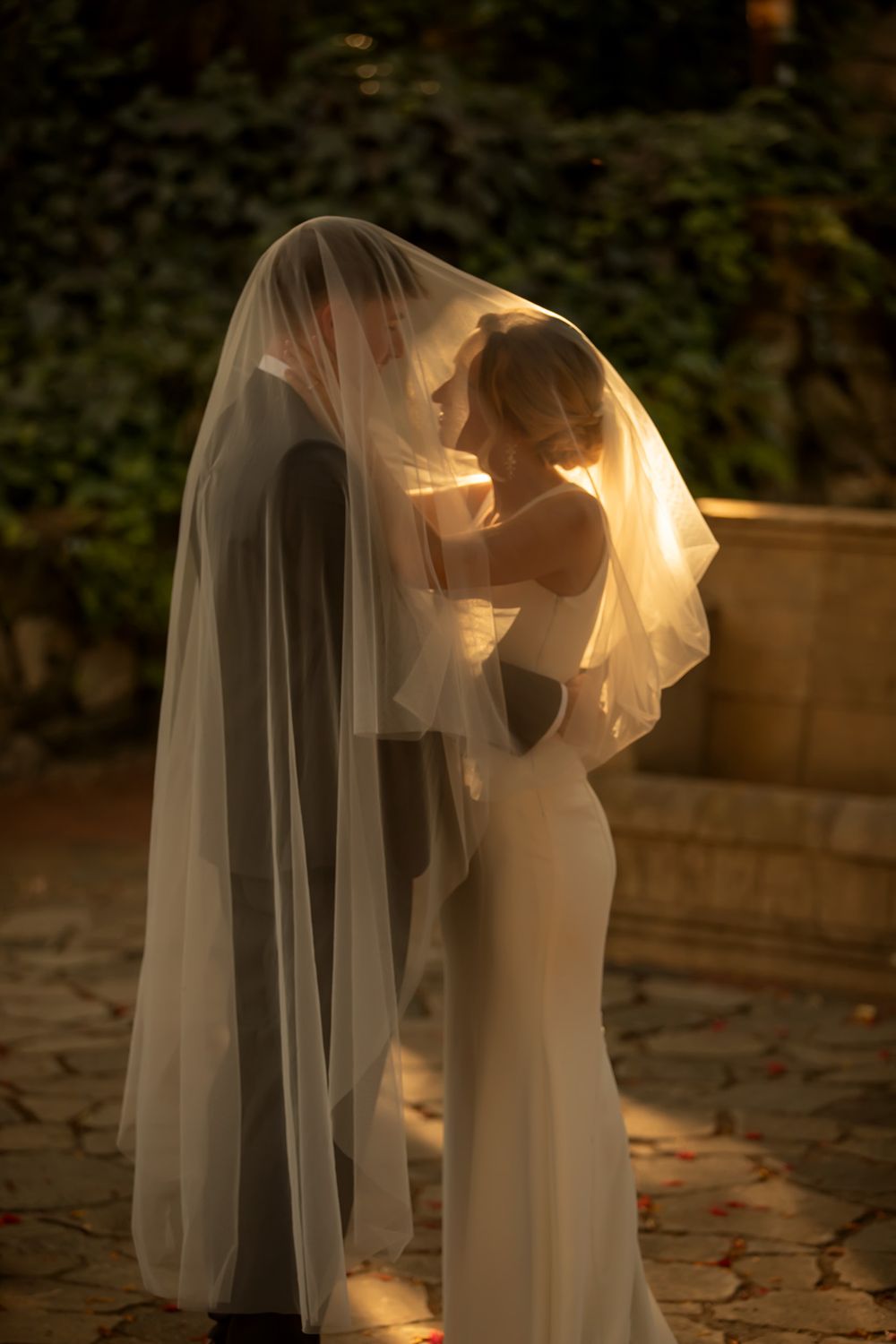 Bride and groom sharing an intimate moment under a flowing white veil in a sunlit garden setting.