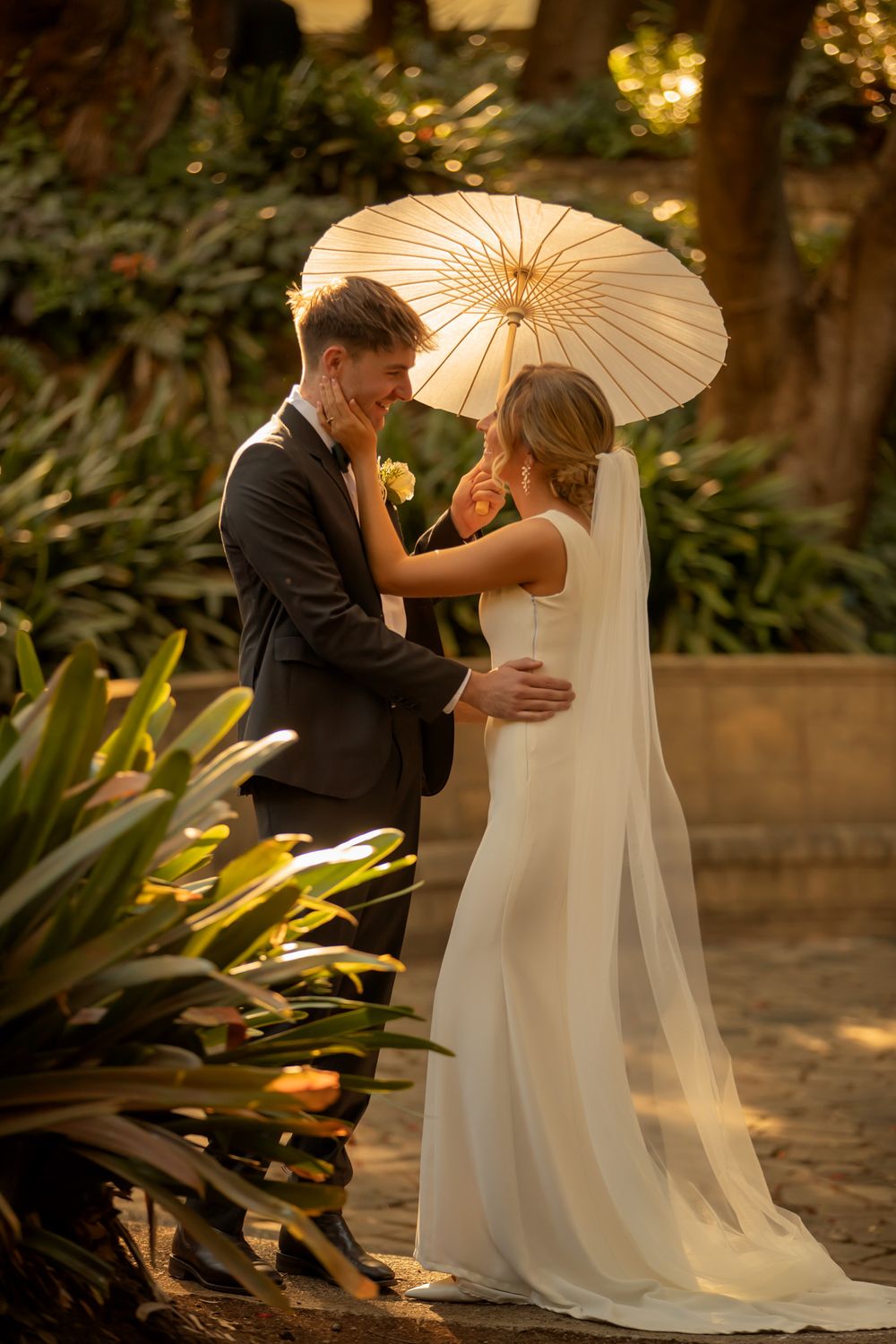 Bride and groom share a romantic moment under a white parasol in a lush garden, golden light illuminating their wedding attire.