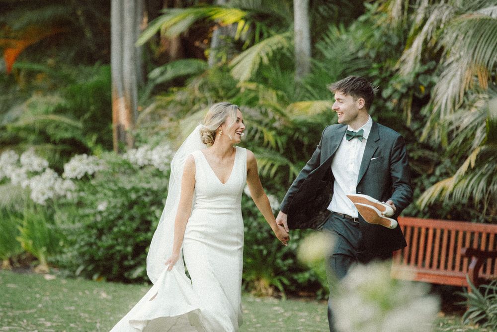 Bride and groom hold hands laughing while walking through lush tropical garden on their wedding day.