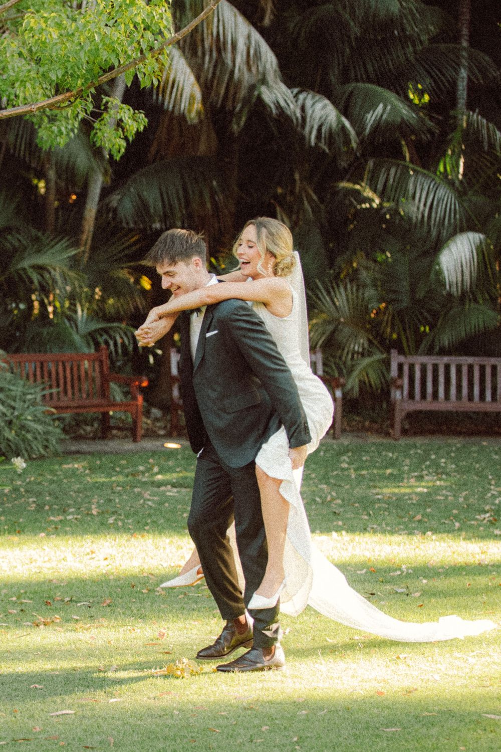 Groom carries laughing bride on his back in sunlit garden with tropical palm trees backdrop.