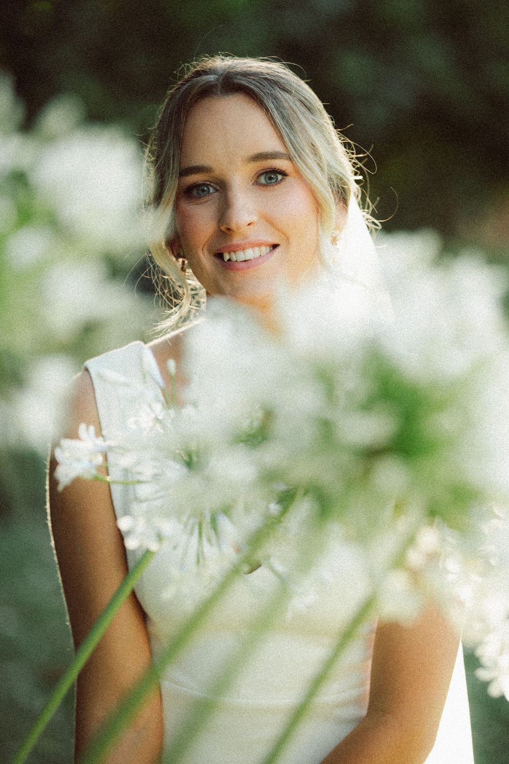 Smiling bride in white dress surrounded by soft-focus white flowers in a lush green garden setting.