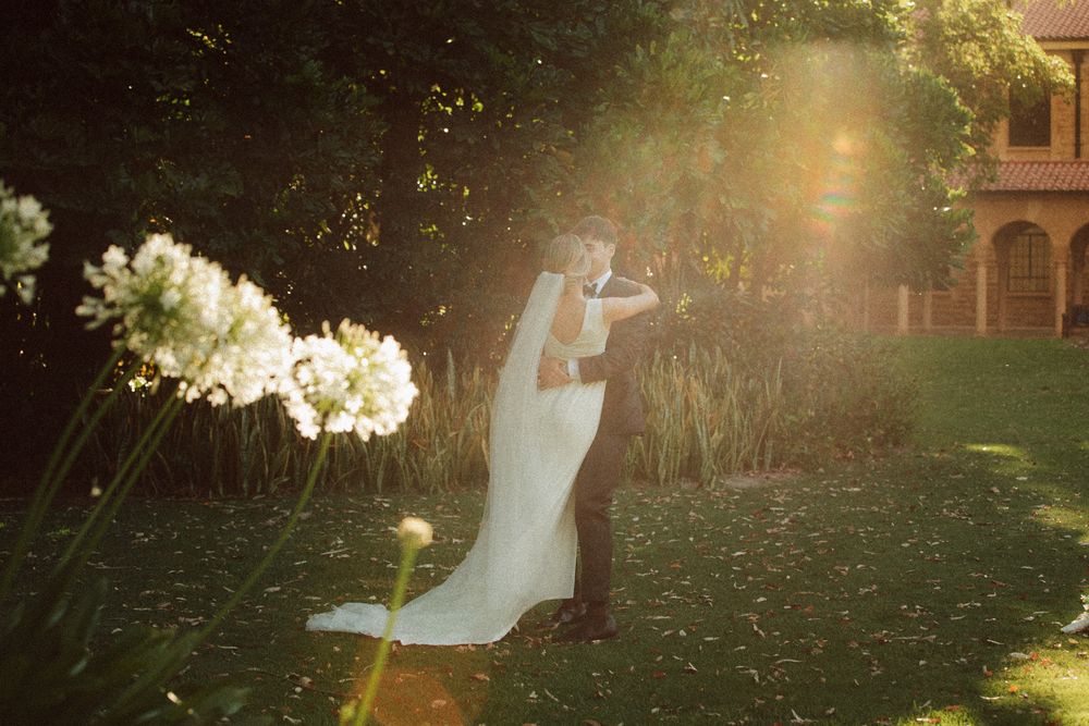 Bride and groom embracing in golden sunlight in a lush garden, with white flowers and warm lens flare creating a romantic atmosphere.