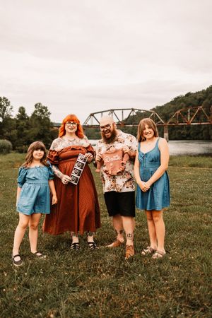 Family of four poses outdoors near a river bridge, holding baby items for a gender reveal photo session.