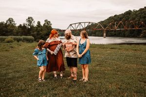 Family of four posing outdoors near a river with a rustic iron bridge backdrop on a grassy field.