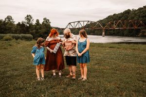 Family gathering closely together on green grass near a scenic river and historic iron bridge.