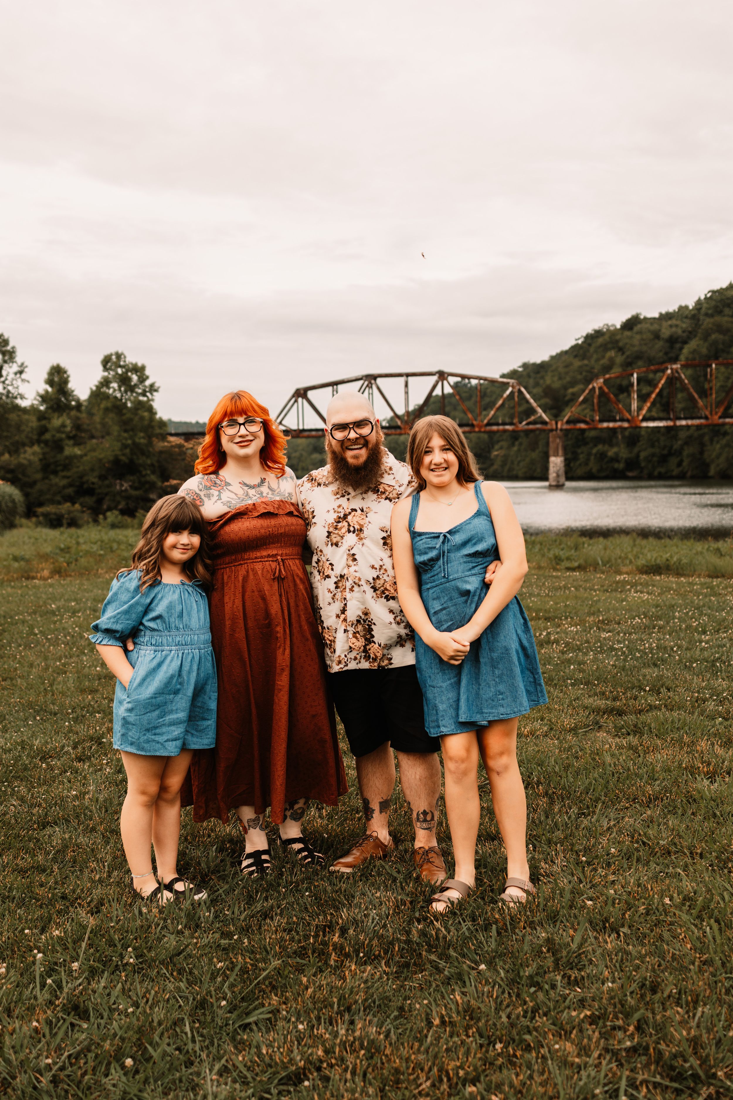 Family of four posing outdoors near a river with a rustic iron bridge backdrop on a cloudy day.