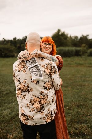 A couple embraces outdoors, woman with red hair smiling, man holding ultrasound photos on his back in a floral shirt.