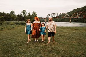 Family of four walking in a grassy field near a river with a rustic bridge in the background.