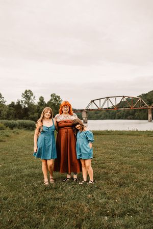 A woman in a rust dress with two girls in blue dresses pose on a grassy riverbank with a steel bridge behind them.