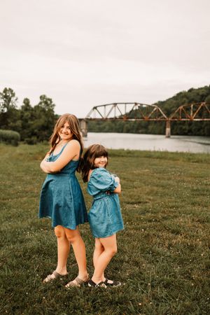 Two girls in matching blue denim dresses stand back-to-back on grass near a river with a rustic bridge backdrop.