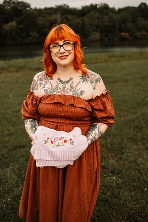 Smiling redhead woman with tattoos wearing rust off-shoulder dress holds embroidered pouch outdoors.