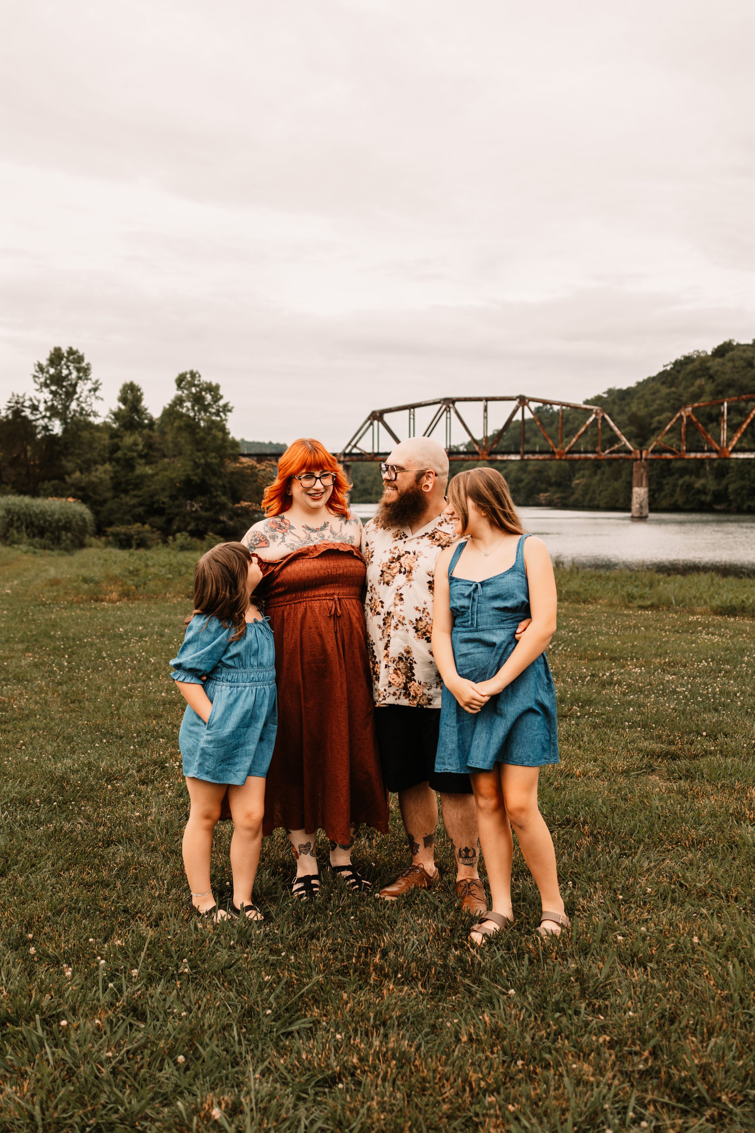 Family of four posing outdoors near a river and bridge, with two girls in blue dresses and parents in warm tones.