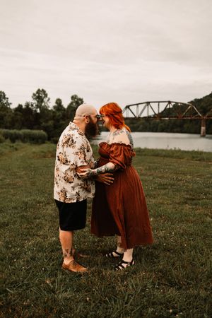 A bald bearded man and red-haired woman in rust dress embrace on grass by a river bridge.