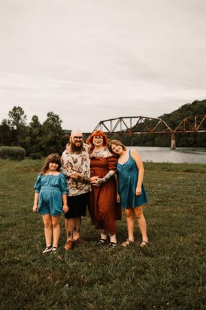Family of four laughing and embracing on green grass near a scenic river and historic iron bridge.