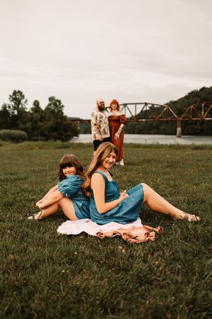 Two girls sitting on grass in teal dresses with parents standing behind near a river bridge.