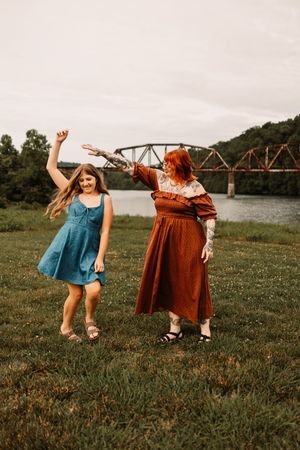 Two women dancing outdoors near a river with a historic bridge, one in a blue dress, the other in a rust-colored maxi dress.