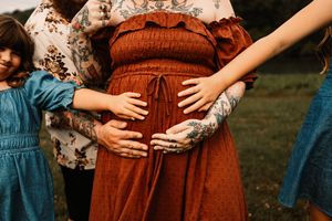 Family members place hands on pregnant womans baby bump in a touching outdoor maternity photo.