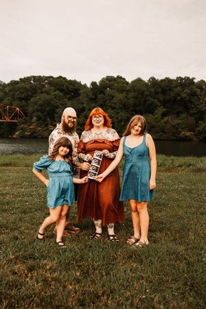 Family of four posing outdoors near a river, with two girls in blue dresses and parents in rustic attire.