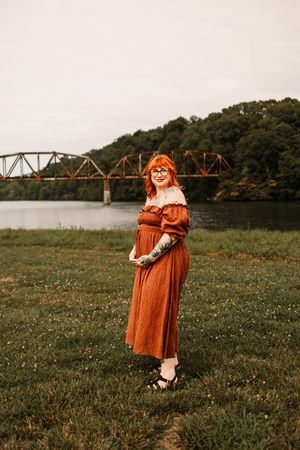 Red-haired woman in rust off-shoulder dress stands by river with iron bridge backdrop on overcast day.