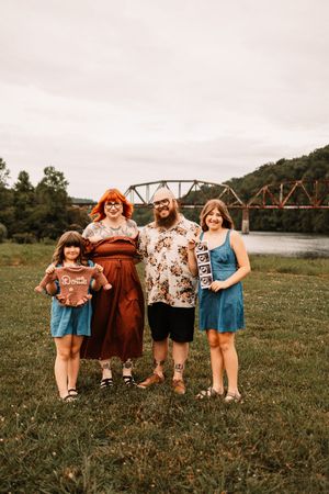 Smiling family of four holding photos outdoors by a river with an old iron bridge in background.