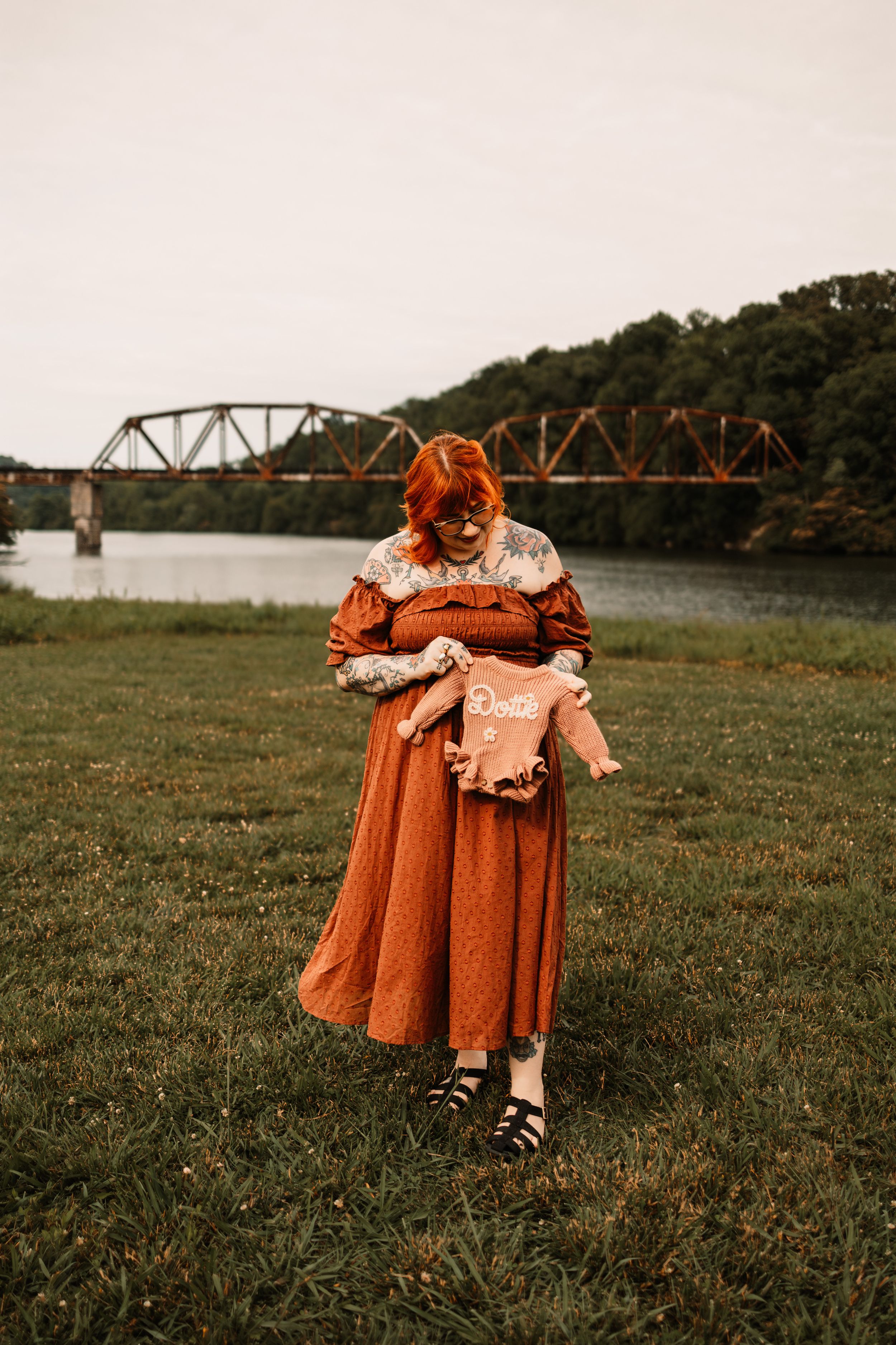 A red-haired pregnant woman in rust dress holds a baby onesie near a river bridge.