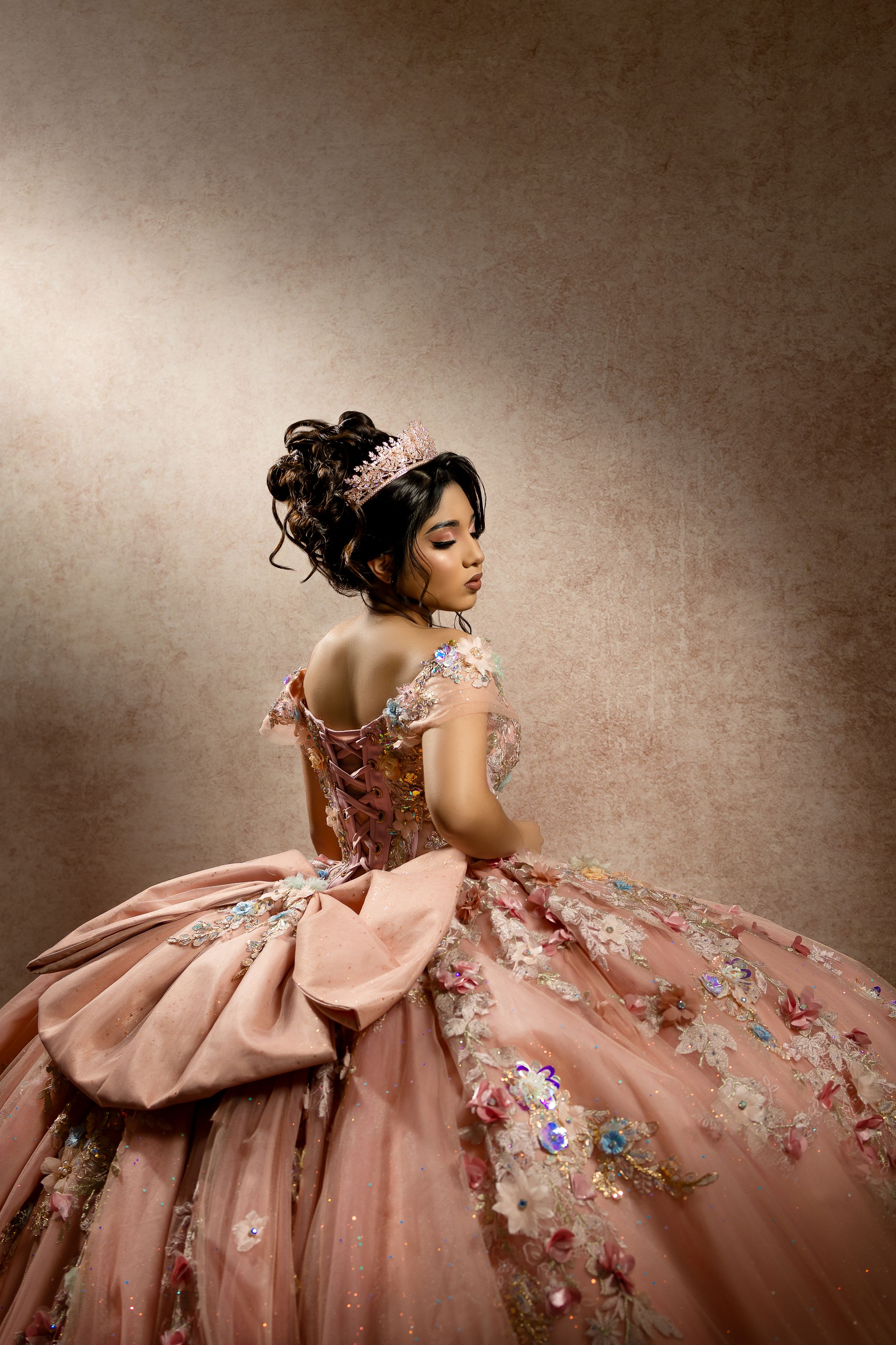 Young woman in pink floral ball gown with tiara poses gracefully, showcasing intricate embroidery and elegant updo hairstyle.