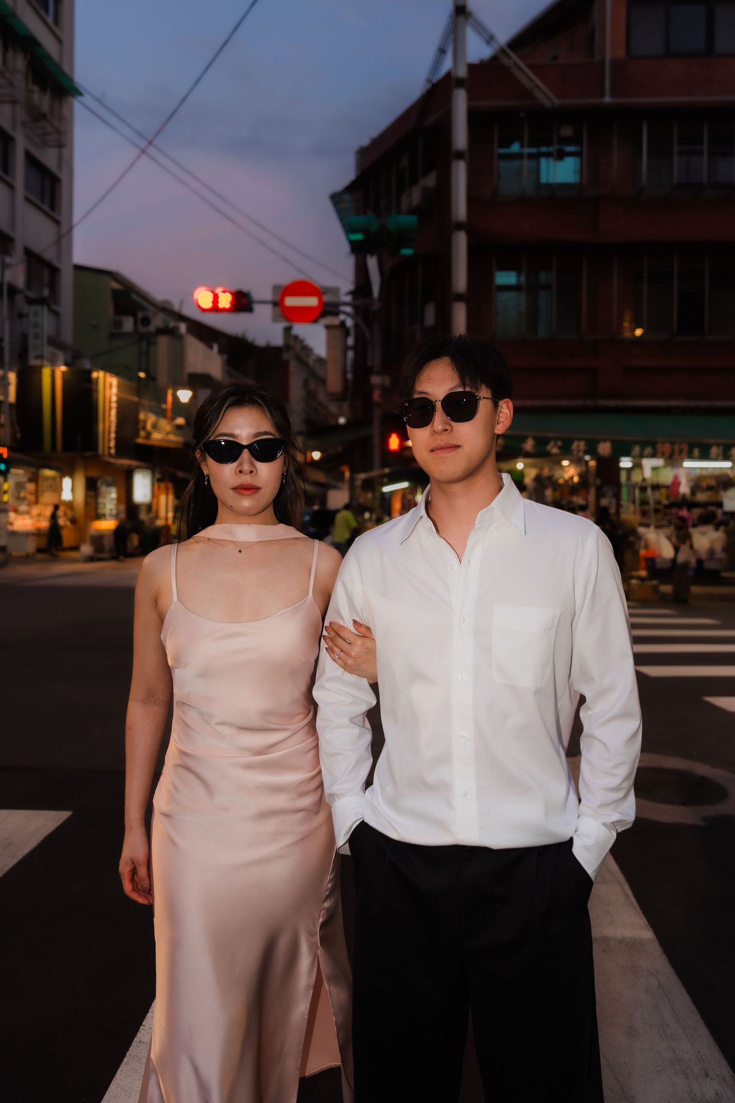 A stylish couple in sunglasses poses on a crosswalk at dusk, woman in satin slip dress, man in white shirt.