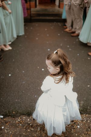 Young flower girl in white dress standing on confetti-covered path flanked by wedding guests.
