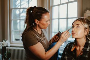Makeup artist applying foundation to bride's face near bright window during wedding preparations.