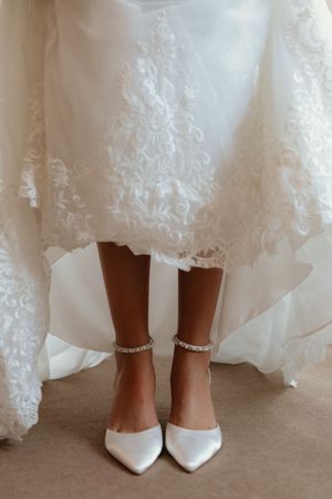 Bride wearing white satin pointed heels with crystal ankle straps beneath lace wedding gown.