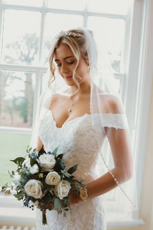 Bride in lace off-shoulder wedding dress holding white rose bouquet standing by bright window with veil.