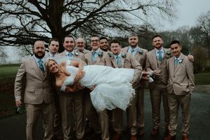 Groomsmen in tan suits with teal ties lifting the bride in a white lace gown outdoors at a wedding.