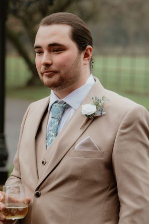 Groom in tan three-piece suit with teal tie and white boutonniere holding a whiskey glass.