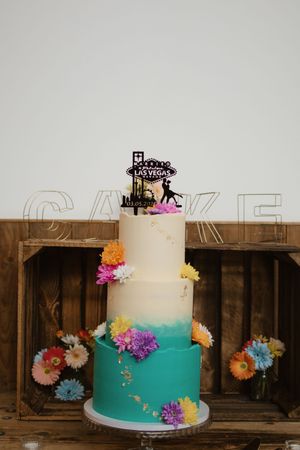 Three-tier Las Vegas themed wedding cake with colorful flowers and teal ombre frosting.
