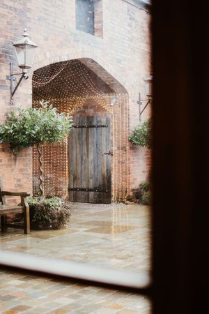 Rustic brick courtyard with wooden door framed by fairy lights viewed through window.