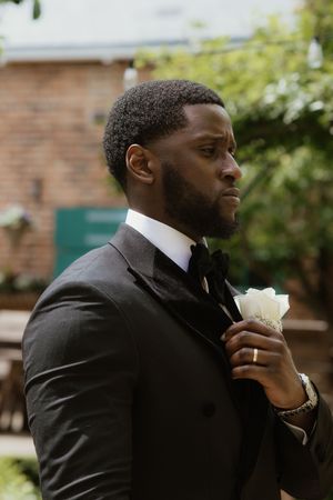 Groom in black tuxedo adjusting white rose boutonniere outdoors.