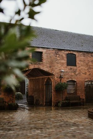 Rustic red brick barn wedding venue with cobblestone courtyard, fairy light canopy entrance, and wooden doors on a rainy day.