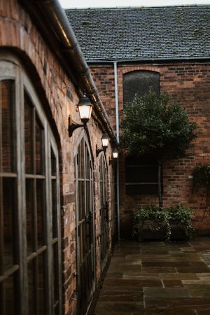 Elegant brick barn venue exterior with arched windows, glowing wall lanterns, and manicured topiary trees in courtyard.