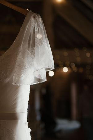 Delicate lace wedding dress and veil hanging in a dimly lit barn with warm bokeh fairy lights in background.