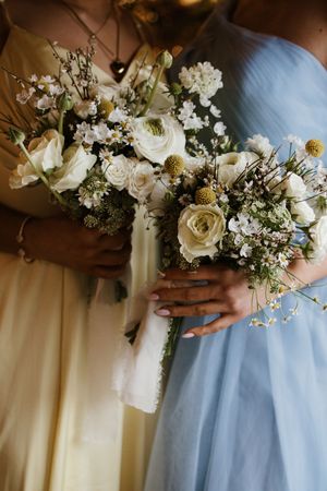 Two bridesmaids in blue and yellow gowns holding wildflower bouquets with white roses and scabiosa blooms.